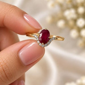 May include: A gold ring featuring a central oval-cut deep red ruby gemstone. The band is accented with a curved row of small, round, clear white diamonds set along the base of the ruby, creating a delicate, contoured design on a polished gold band.