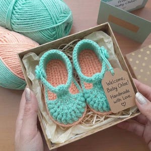 May include: A pair of handmade crochet baby shoes in mint green and peach, featuring a T-strap design with a small button. The shoes are nestled in a gift box with a brown paper tag that reads, Welcome, Baby Chloe! Handmade with Love.