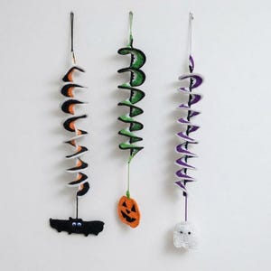 May include: Three hanging crochet spiral decorations featuring Halloween themes. The left spiral is orange, black, and white with a black bat. The center spiral is green and black with an orange pumpkin. The right spiral is purple and white with a white ghost.