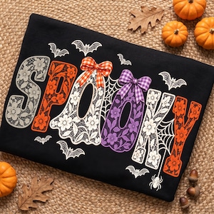 Op de afbeelding: Een zwart t-shirt met het woord SPOOKY in grote letters met een bloemrijk kantpatroon. De letters zijn gekleurd in lichtgrijs, oranje en paars, geaccentueerd met oranje en paarse geruite strikken, spinnenwebben en kleine witte vleermuizen.