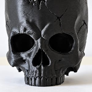 May include: A matte black skull-shaped container with a jagged, crown-like top edge. The surface features realistic anatomical details including deep eye sockets, a nasal cavity, and a row of teeth, with visible cracks across the forehead area.