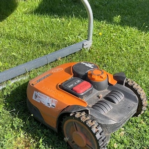 Könnte beinhalten: Ein orange-schwarzer Mähroboter mit einem markanten roten Stoppknopf auf dem oberen Bedienfeld, einem runden Einstellrad und profilierten Rädern, der auf einer grünen Rasenfläche neben einem gebogenen Metallrahmen steht.