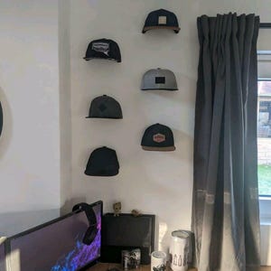 Könnte beinhalten: Eine Sammlung von sieben Baseballkappen, die an einer weißen Wand befestigt sind. Die Kappen variieren in der Farbe, darunter Schwarz, Grau und Marineblau, mit flachen oder gebogenen Schirmen. Einige Kappen zeigen Logos oder Aufnäher auf der Vorderseite.