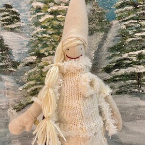 Op de afbeelding: Een witte vilten pop die een witte gebreide trui en een witte vilten hoed met een witte bontkraag draagt. De pop heeft een wit vilten gezicht met een rode glimlach en zwarte ogen. De pop zit in een besneeuwd winterlandschap.