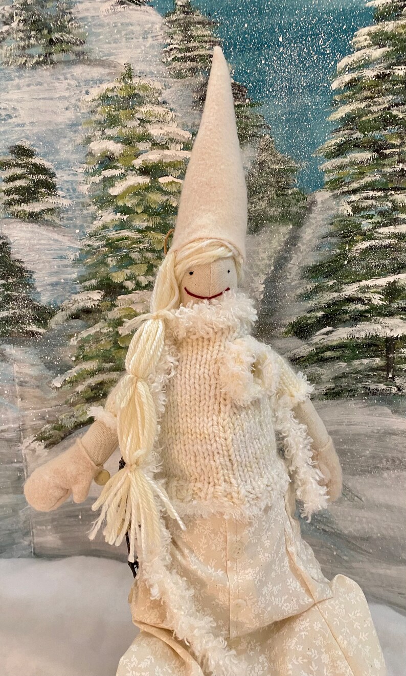 Op de afbeelding: Een witte vilten pop die een witte gebreide trui en een witte vilten hoed met een witte bontkraag draagt. De pop heeft een wit vilten gezicht met een rode glimlach en zwarte ogen. De pop zit in een besneeuwd winterlandschap.