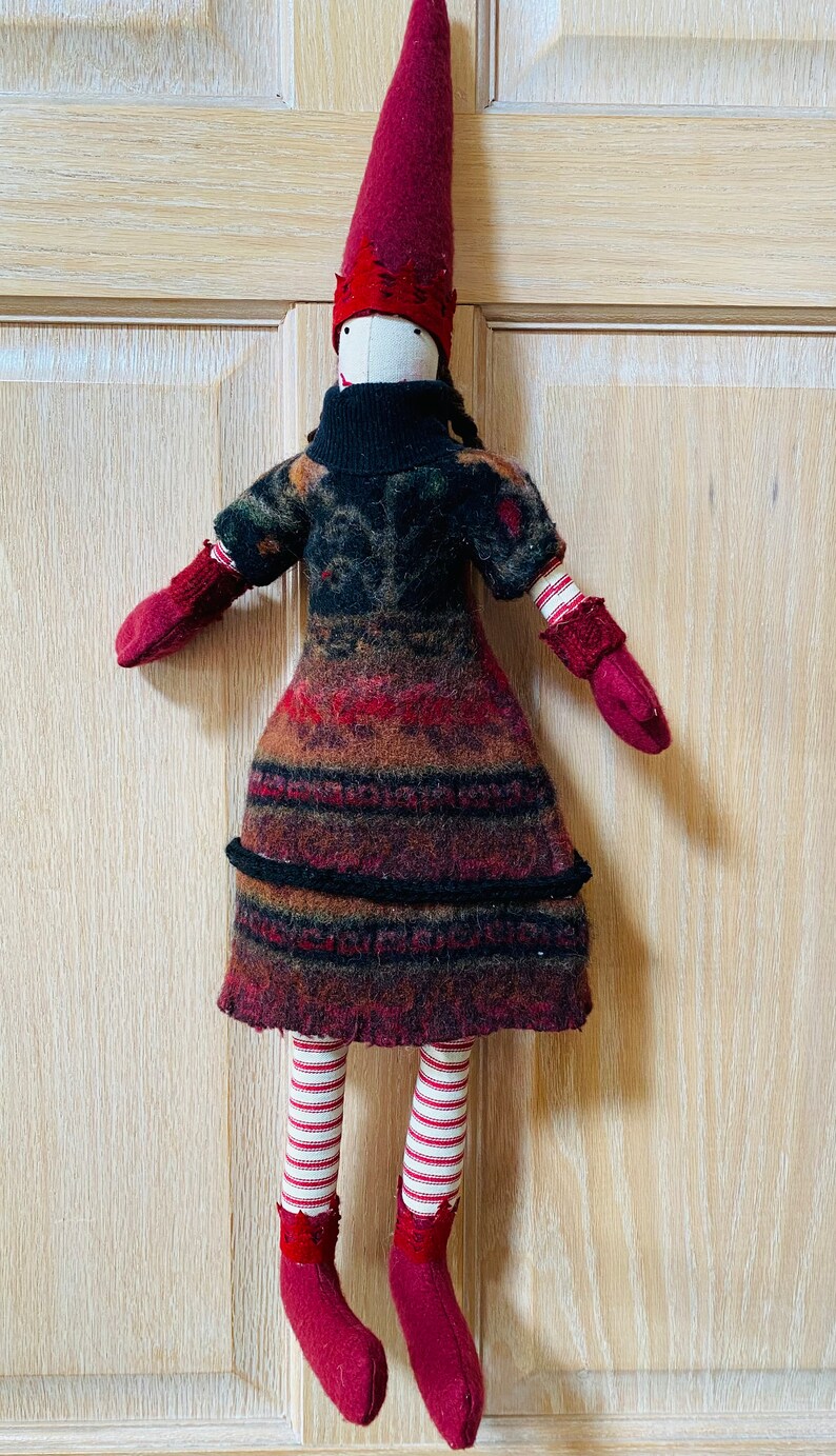 Peut inclure: Poup&eacute;e artisanale avec un grand chapeau pointu bordeaux et des mitaines et bottes assorties. La poup&eacute;e a un col roul&eacute; noir et une robe &agrave; motifs dans les tons de rouge, marron et noir. Les jambes de la poup&eacute;e sont ray&eacute;es rouge et blanc.