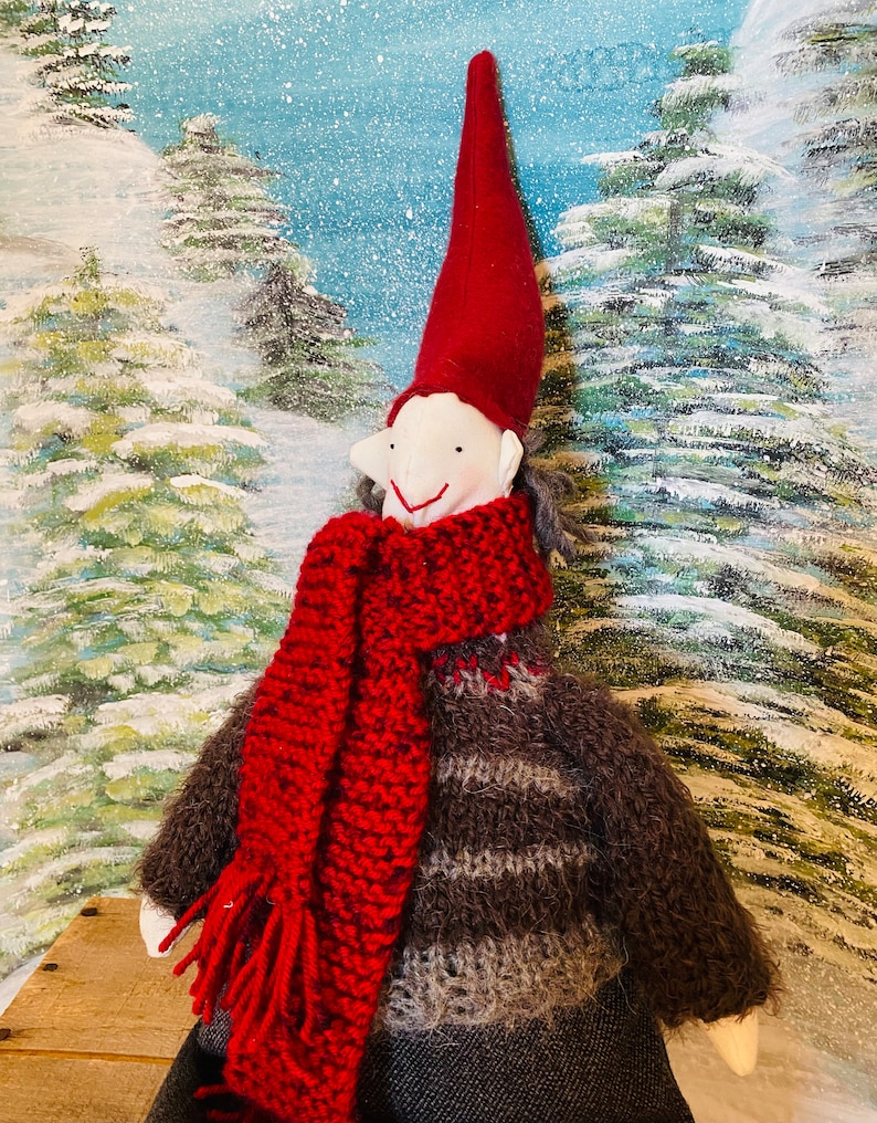 May include: A handmade Christmas gnome doll with a red pointed hat and scarf. The gnome wears a brown and grey sweater and a dark skirt. The background features a winter scene with snow-covered trees.