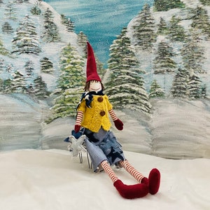 May include: A handmade doll with a red hat, yellow sweater, blue jeans, and red and white striped legs. The doll is holding a white star and sitting in a snowy winter scene.