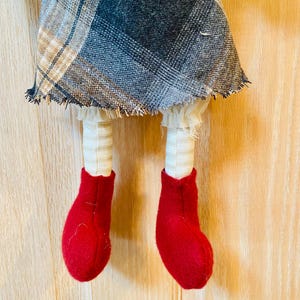 May include: A handmade rag doll with a plaid skirt, white ruffles, and red felt boots. The doll is hanging from a wooden surface.
