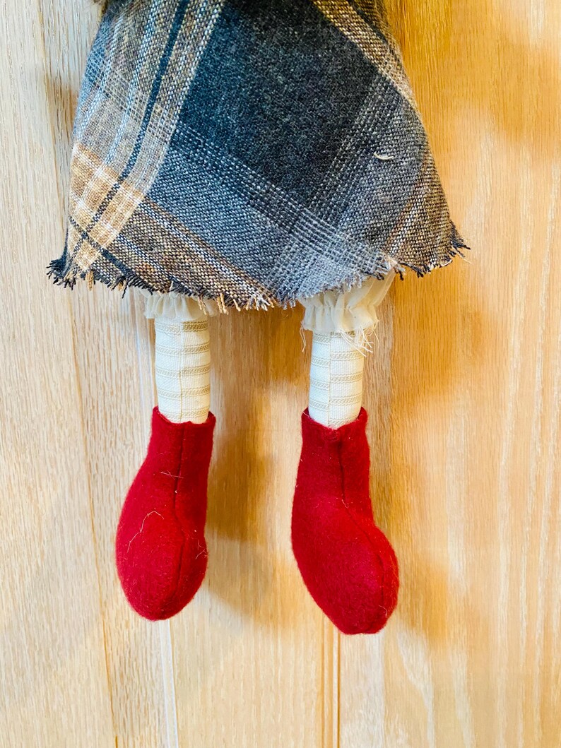 May include: A handmade rag doll with a plaid skirt, white ruffles, and red felt boots. The doll is hanging from a wooden surface.