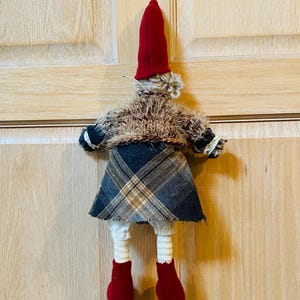 May include: A handmade gnome doll with a red hat, brown fur coat, plaid skirt, and red boots. The gnome is hanging on a wooden door.