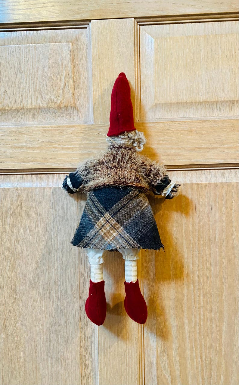 May include: A handmade gnome doll with a red hat, brown fur coat, plaid skirt, and red boots. The gnome is hanging on a wooden door.