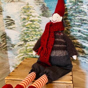 May include: A festive Christmas elf doll with a red pointed hat, scarf, and heart-shaped feet. The elf is dressed in a striped sweater and sits on a wooden crate against a snowy forest backdrop. The doll's legs are striped in red and white.