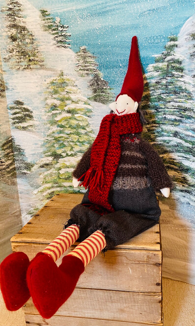 May include: A festive Christmas elf doll with a red pointed hat, scarf, and heart-shaped feet. The elf is dressed in a striped sweater and sits on a wooden crate against a snowy forest backdrop. The doll's legs are striped in red and white.