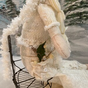 Op de afbeelding: Een witte pop met een gebreide trui en een witte rok zit op een draadstoel. De pop heeft lang wit haar en een takje groen in haar zak.