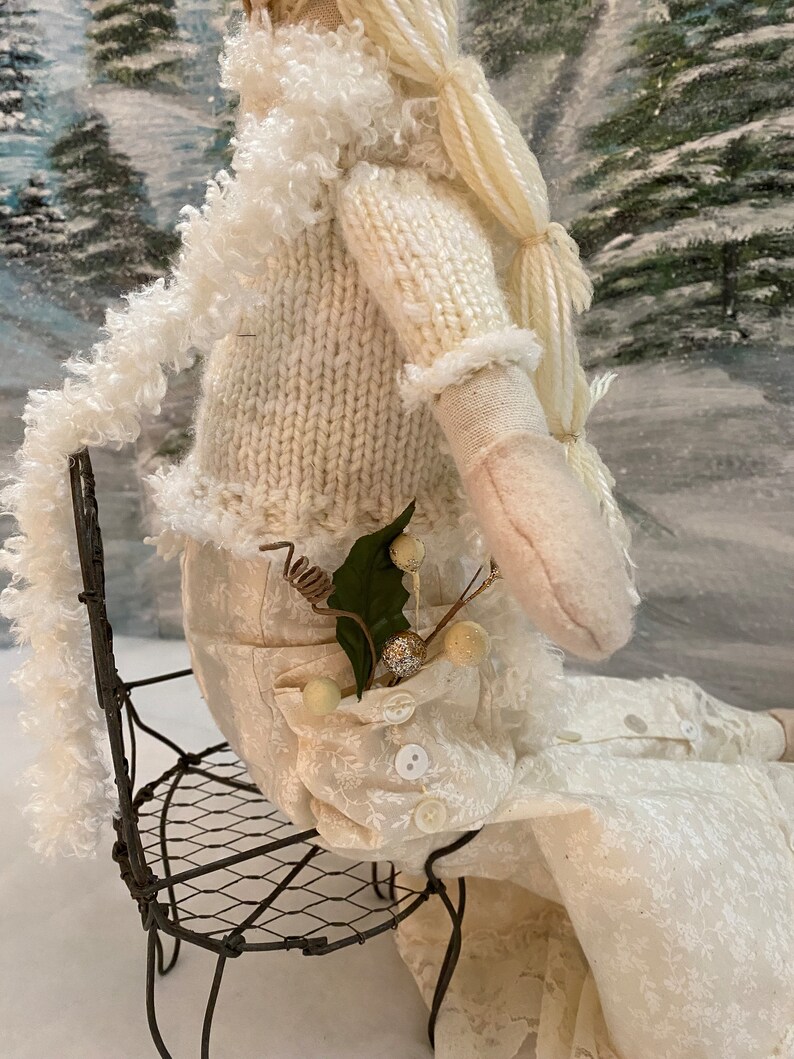 Op de afbeelding: Een witte pop met een gebreide trui en een witte rok zit op een draadstoel. De pop heeft lang wit haar en een takje groen in haar zak.