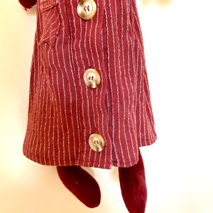 Op de afbeelding: Een bordeauxrode pop met een verticaal gestreepte jurk met drie grote, ronde, goudkleurige knopen. De jurk heeft een klein zakje aan de linkerkant. De benen en armen van de pop zijn ook bordeauxrood.