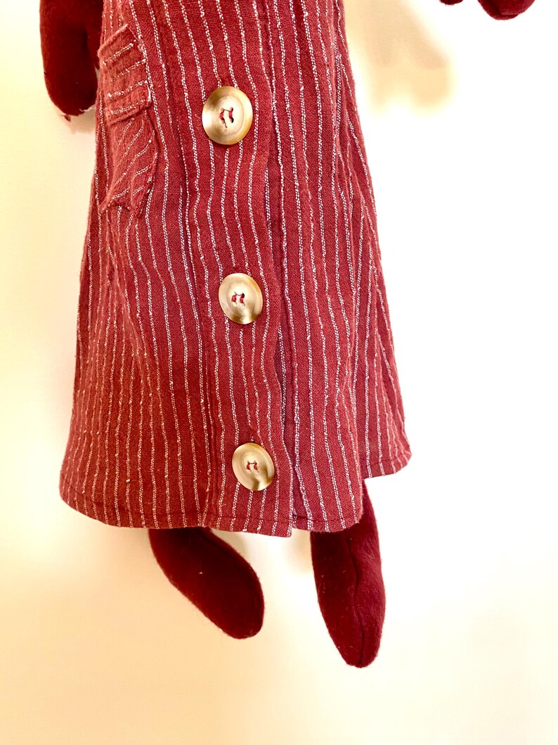 Op de afbeelding: Een bordeauxrode pop met een verticaal gestreepte jurk met drie grote, ronde, goudkleurige knopen. De jurk heeft een klein zakje aan de linkerkant. De benen en armen van de pop zijn ook bordeauxrood.