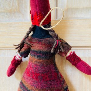 Peut inclure: Poup&eacute;e artisanale avec un grand chapeau en feutre bordeaux et une robe assortie. La poup&eacute;e a des tresses brunes, des moufles rouges et un d&eacute;tail en forme de c&oelig;ur sur la robe. Une ficelle de jute est attach&eacute;e au chapeau pour l'accrocher.