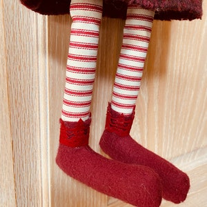 Peut inclure: Une poup&eacute;e faite &agrave; la main avec une jupe bordeaux et des jambes en tissu ray&eacute; rouge et blanc. Les pieds de la poup&eacute;e sont en feutre bordeaux, et la poup&eacute;e est suspendue &agrave; une surface en bois.