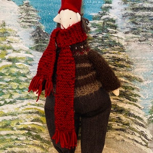 May include: A handmade Christmas gnome doll with a long red hat, red scarf, and dark grey trousers. The doll has a white face and is wearing a brown and grey jumper. The doll's legs are striped red and white, and it has red boots. The background is a winter scene.