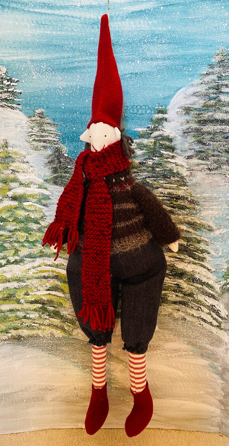 May include: A handmade Christmas gnome doll with a long red hat, red scarf, and dark grey trousers. The doll has a white face and is wearing a brown and grey jumper. The doll's legs are striped red and white, and it has red boots. The background is a winter scene.