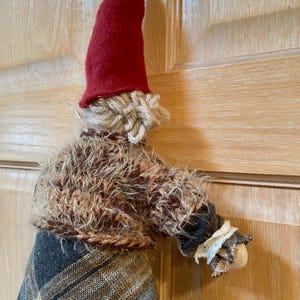 May include: A handmade gnome doll with a red felt hat, brown yarn hair, and a brown and black plaid skirt. The gnome is wearing a brown fuzzy sweater with a white lace trim on the sleeve.