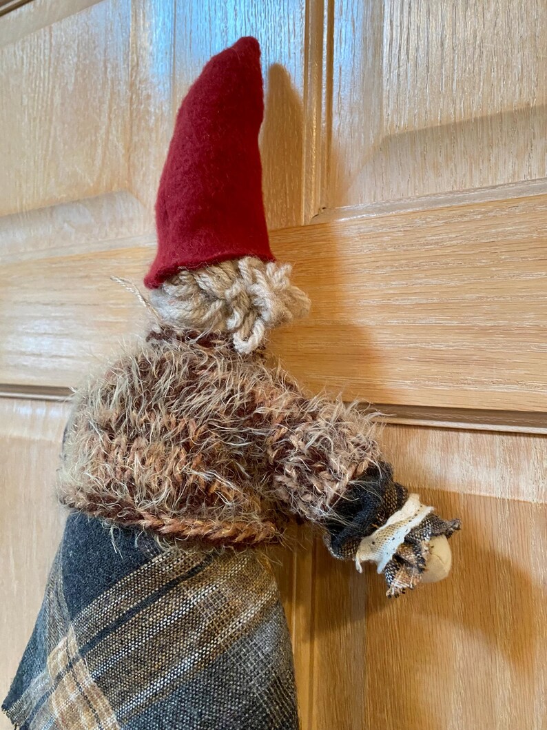 May include: A handmade gnome doll with a red felt hat, brown yarn hair, and a brown and black plaid skirt. The gnome is wearing a brown fuzzy sweater with a white lace trim on the sleeve.