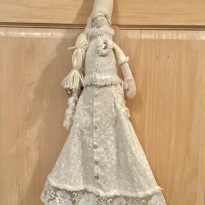 Op de afbeelding: Een witte stoffen pop met een witte gebreide trui, een witte rok met kant en een witte punthoed. De pop hangt aan een houten deur.