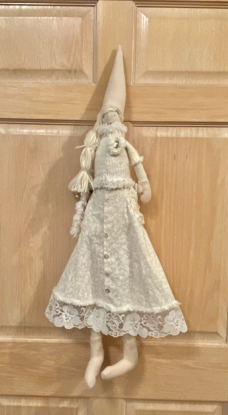 Op de afbeelding: Een witte stoffen pop met een witte gebreide trui, een witte rok met kant en een witte punthoed. De pop hangt aan een houten deur.