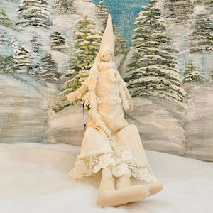 Op de afbeelding: Een witte stoffen pop met een lange witte jurk en een witte hoed. De pop zit op een slee in een besneeuwd winterlandschap.