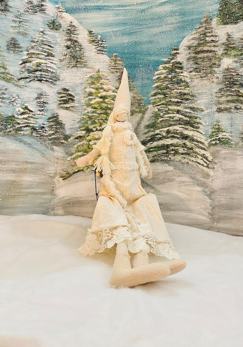 Op de afbeelding: Een witte stoffen pop met een lange witte jurk en een witte hoed. De pop zit op een slee in een besneeuwd winterlandschap.