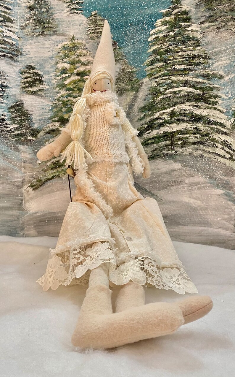 Op de afbeelding: Een witte stoffen pop met een witte gebreide trui en een witte rok met kant. De pop draagt een witte punthoed en heeft een wit stoffen gezicht met geschilderde kenmerken. De pop zit op een wit oppervlak met een besneeuwde achtergrond.