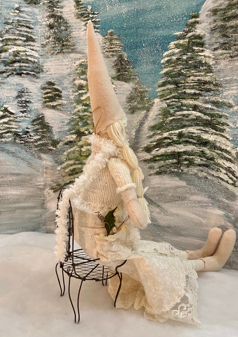 Op de afbeelding: Een witte gnoompup met een lange witte hoed en een witte jurk zit op een draadstoel. De pop houdt een klein boeket groene bladeren en witte bessen vast. De pop zit voor een besneeuwde bos sc&egrave;ne.