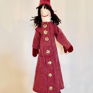 Op de afbeelding: Een handgemaakte pop met een rood-witte gestreepte jas, bijpassende hoed en wanten. De pop heeft zwart garen haar en een lachend gezicht. De jas is aan de voorkant dichtgeknoopt met grote, ronde knopen. De pop hangt aan een touwtje.