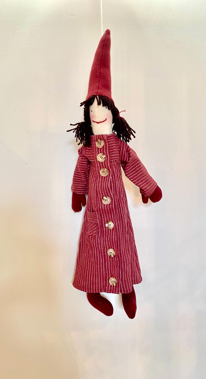 Op de afbeelding: Een handgemaakte pop met een rood-witte gestreepte jas, bijpassende hoed en wanten. De pop heeft zwart garen haar en een lachend gezicht. De jas is aan de voorkant dichtgeknoopt met grote, ronde knopen. De pop hangt aan een touwtje.