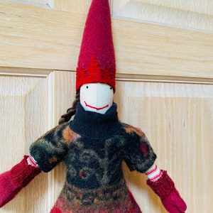 Peut inclure: Poup&eacute;e artisanale avec un long chapeau pointu bordeaux et une robe &agrave; motifs. La poup&eacute;e a un visage blanc avec un simple sourire et des cheveux bruns tress&eacute;s. Ses mitaines et la garniture du chapeau sont bordeaux, et la robe pr&eacute;sente un m&eacute;lange de couleurs.