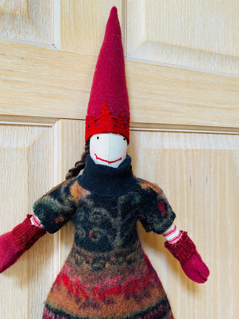 Peut inclure: Poup&eacute;e artisanale avec un long chapeau pointu bordeaux et une robe &agrave; motifs. La poup&eacute;e a un visage blanc avec un simple sourire et des cheveux bruns tress&eacute;s. Ses mitaines et la garniture du chapeau sont bordeaux, et la robe pr&eacute;sente un m&eacute;lange de couleurs.