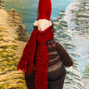 May include: A handmade Christmas gnome doll with a red pointed hat, a red knitted scarf, and a brown and grey jumper. The doll has a white face and is wearing dark grey trousers and striped socks. The background is a winter scene.