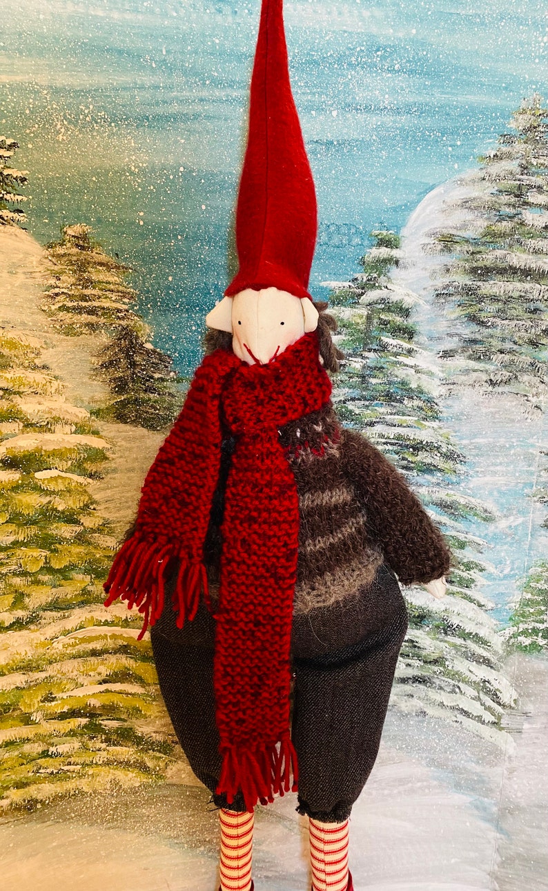 May include: A handmade Christmas gnome doll with a red pointed hat, a red knitted scarf, and a brown and grey jumper. The doll has a white face and is wearing dark grey trousers and striped socks. The background is a winter scene.