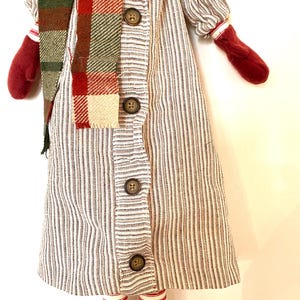 May include: A handmade doll in a striped dress with button details, a plaid scarf, and red mittens. The doll has red and white striped legs and red shoes. The doll is a festive holiday decoration.