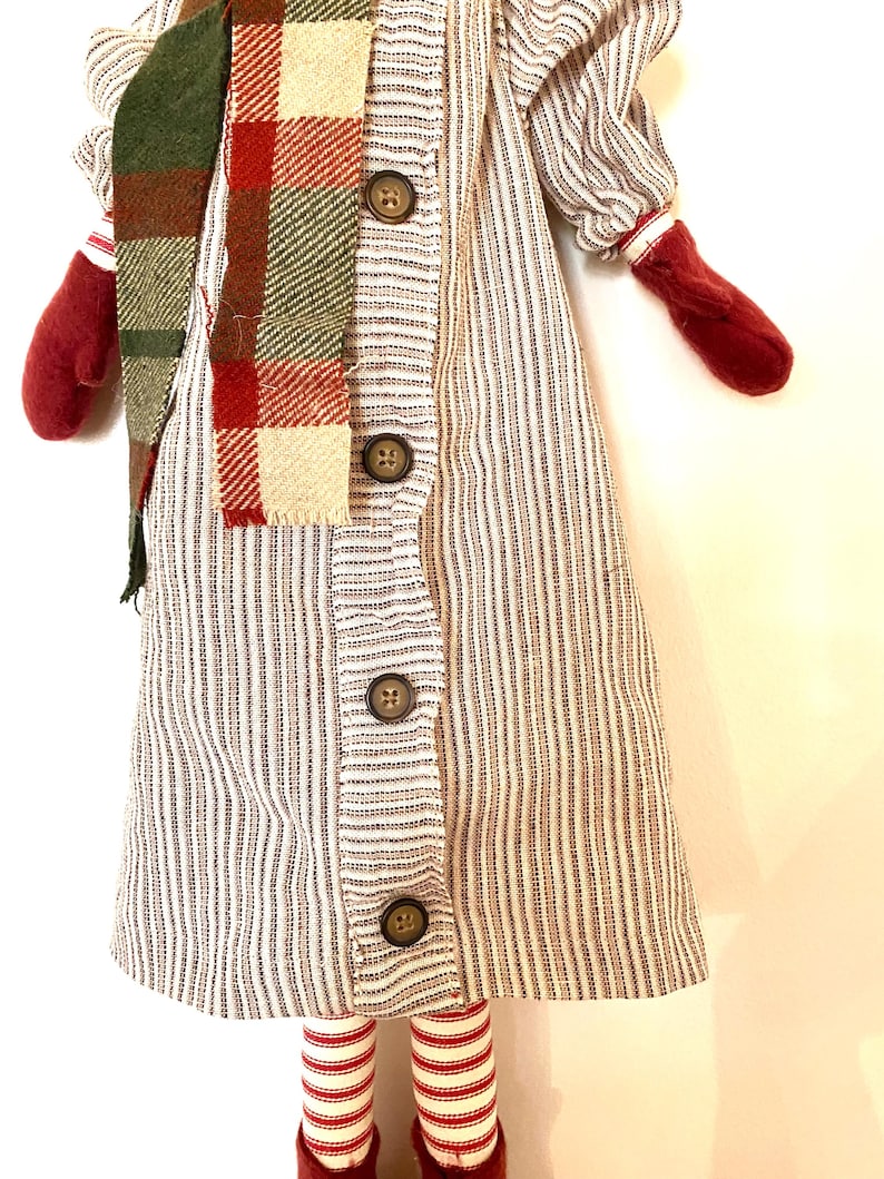 May include: A handmade doll in a striped dress with button details, a plaid scarf, and red mittens. The doll has red and white striped legs and red shoes. The doll is a festive holiday decoration.