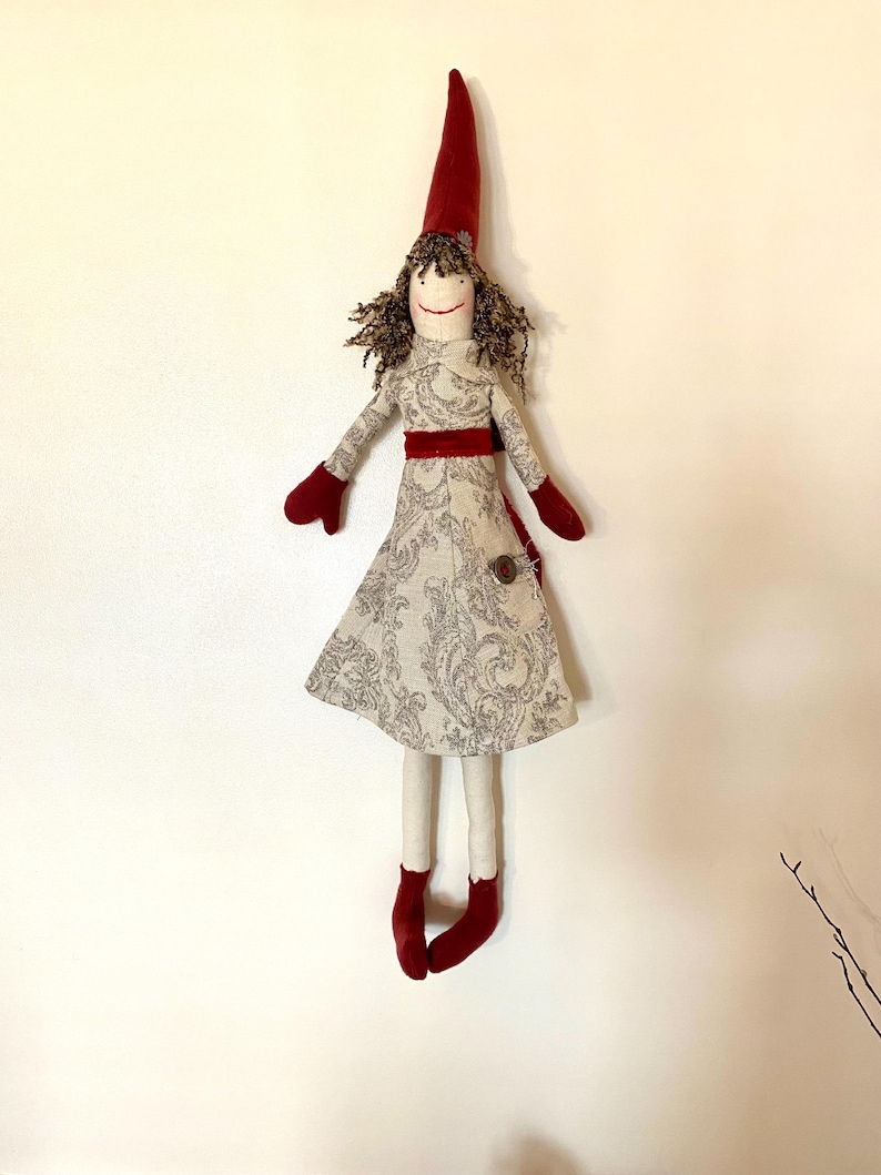 以下が含まれることがあります： 赤いとんがり帽子、お揃いのミトン、靴を履いた手作りの人形。人形のドレスは、グレーの花柄と赤いサッシュが付いた明るい色です。人形は茶色の巻き毛で、笑顔の顔をしています。