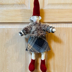 May include: A handmade felt doll with a red hat, brown fur coat, plaid skirt, and red shoes. The doll is hanging on a wooden door.