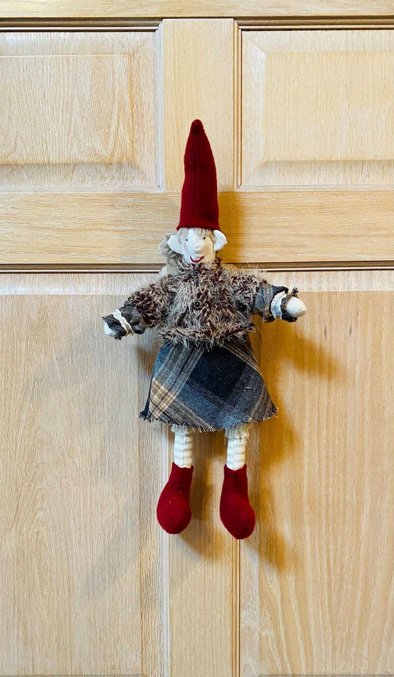 May include: A handmade felt doll with a red hat, brown fur coat, plaid skirt, and red shoes. The doll is hanging on a wooden door.