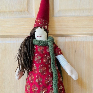 May include: A handmade doll with long brown hair, wearing a red dress with gold sequins, a green scarf, and a red hat.