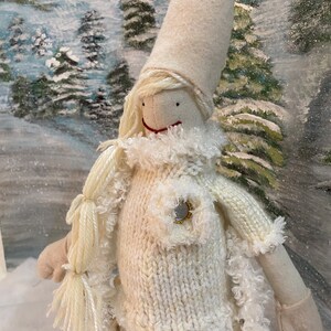 Op de afbeelding: Een witte gebreide pop met een witte bontkraag en een witte muts. De pop heeft een rond gezicht met zwarte ogen en een rode mond. De pop draagt een witte gebreide trui met een gouden knoop. De pop staat voor een besneeuwde achtergrond.