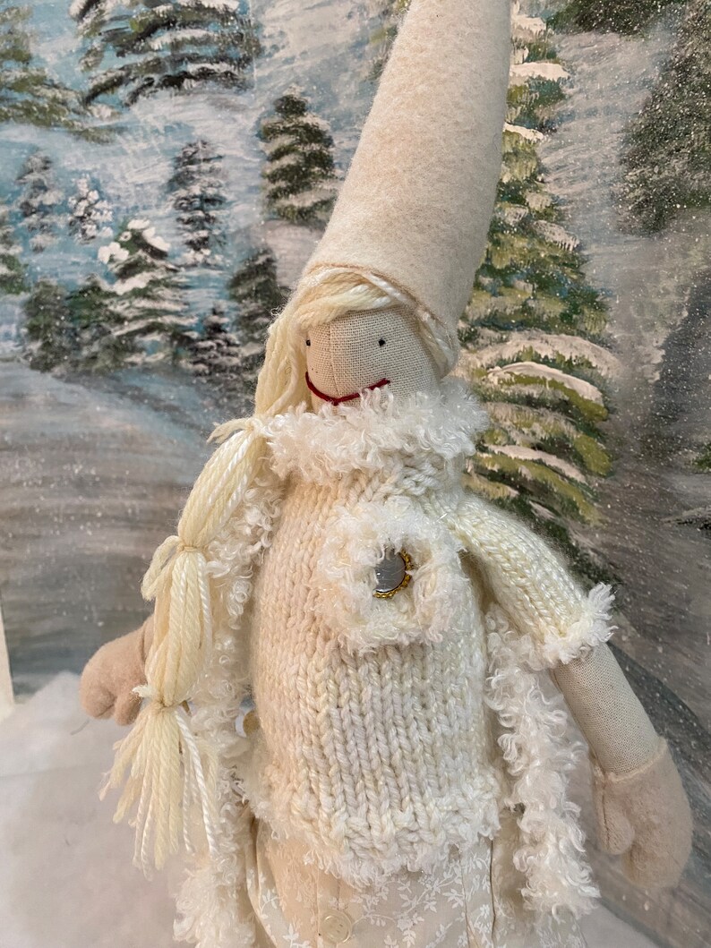 Op de afbeelding: Een witte gebreide pop met een witte bontkraag en een witte muts. De pop heeft een rond gezicht met zwarte ogen en een rode mond. De pop draagt een witte gebreide trui met een gouden knoop. De pop staat voor een besneeuwde achtergrond.