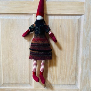 May include: A handmade fabric doll with a long, pointed burgundy hat and matching mittens and shoes. The doll wears a patterned dress in shades of red, brown, and black, with a black collar. Its legs are striped red and white. The doll is hanging on a wooden door.