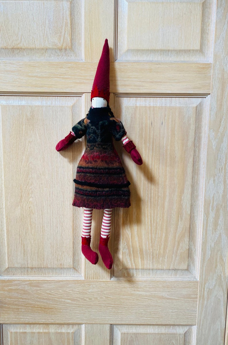 Peut inclure: Poup&eacute;e en tissu faite &agrave; la main avec un long chapeau pointu bordeaux et des mitaines et chaussures assorties. La poup&eacute;e porte une robe &agrave; motifs dans les tons de rouge, marron et noir, avec un col noir. Ses jambes sont ray&eacute;es rouge et blanc. La poup&eacute;e est suspendue &agrave; une porte en bois.